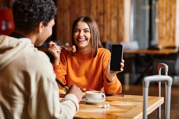 Glückliche junge Frau in orangefarbenem Pullover zeigt ihrem schwarzen Freund während des Essens fröhlich ihr Handy — Stockfoto