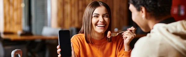Mujer feliz en suéter naranja felizmente mostrando su teléfono al novio negro durante la comida, pancarta - foto de stock