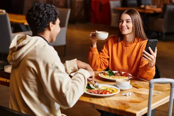 Glückliche Frau in orangefarbenem Pullover, die während des Essens ihr Handy und ihre Kaffeetasse neben ihrem schwarzen Freund hält — Stockfoto