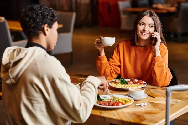Relaxed woman having pleasant phone call while holding a cup of coffee near black boyfriend in cafe — Stock Photo