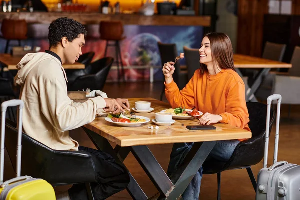 Diverse couple sharing a joyful moment over a healthy meal in a trendy cafe, travel luggage — Stock Photo