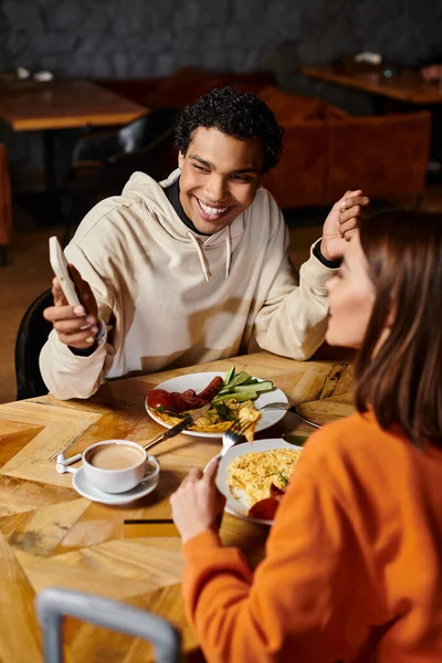 Пара, насолоджуючись затишною їжею разом, чорний чоловік показує смартфон дівчині — стокове фото