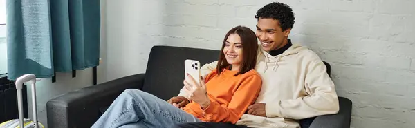 Heureux couple diversifié partage un moment confortable, défilant à travers les téléphones sur un canapé confortable, bannière — Photo de stock