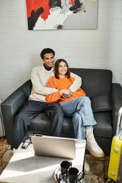 Happy diverse couple cozily hugging on a couch in their stylish living room near laptop on table — Stock Photo