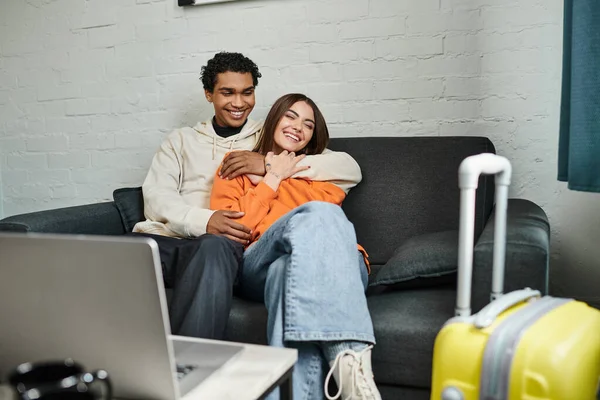 Щаслива багатоетнічна пара затишно обіймається на дивані у своїй стильній вітальні біля ноутбука на столі — стокове фото
