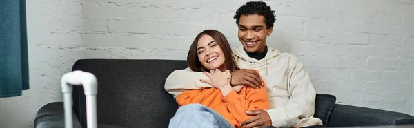 Счастливая многонациональная пара уютно обнимая на диване в стильной гостиной общежития, баннер — стоковое фото