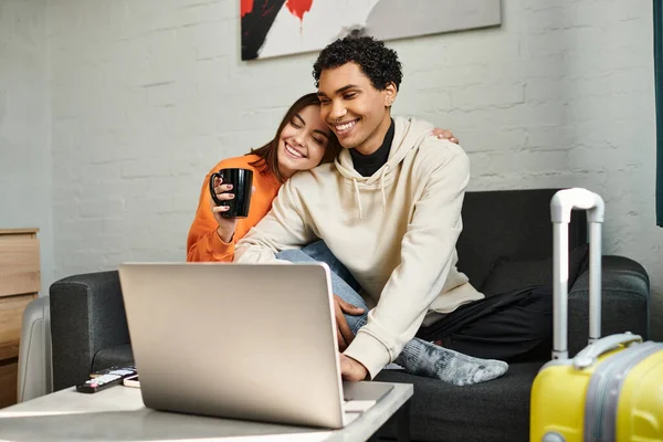 Happy diverse couple shares a cozy moment as they sit on couch and using laptop, hostel — Stock Photo