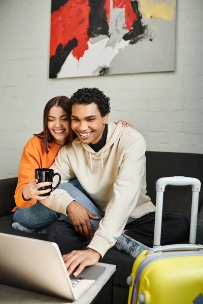Excited interracial couple shares a cozy moment as they sit on couch and using laptop, hostel — Stock Photo