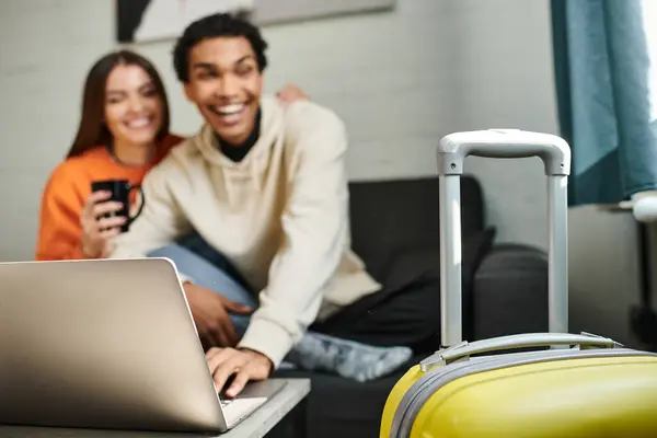 Focus on yellow luggage, happy diverse couple traveling together hostel living room and laptop — Stock Photo