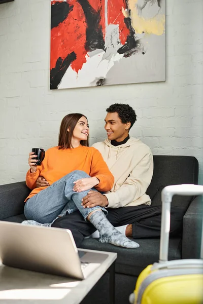 Happy interracial couple sitting on a couch in hostel, surrounded by laptop and luggage — Stock Photo