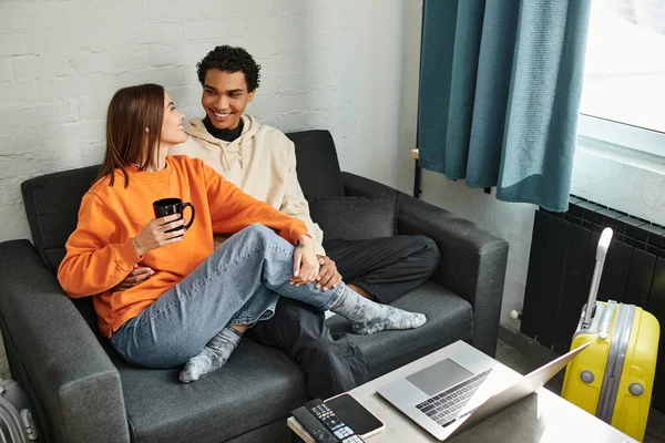 Happy diverse couple in cozy clothing sitting on a couch in hostel, surrounded by laptop and luggage — Stock Photo