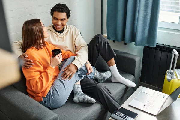 Feliz pareja diversa sentado en un sofá en el albergue, rodeado de aparatos y equipaje - foto de stock
