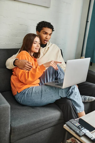 Paar sitzt auf Couch und surft Laptop, Frau zeigt auf Bildschirm neben schwarzem Freund — Stockfoto