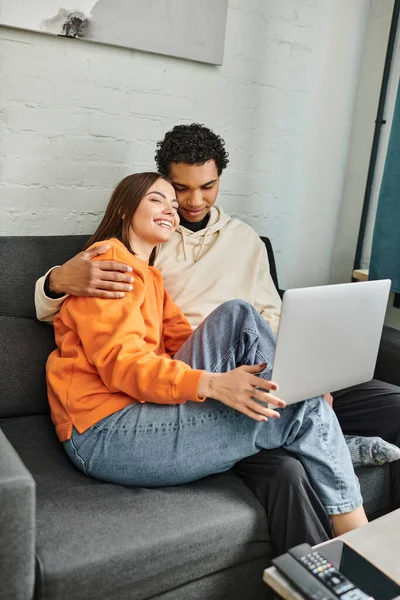 Happy diverse couple snuggle on couch while watching movie on laptop, hostel room — Stock Photo