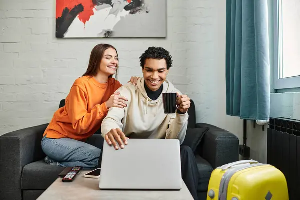 Excited diverse couple seated on couch with a laptop, possibly booking a trip in hostel room — Stock Photo