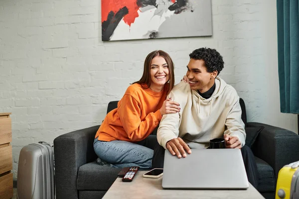 Feliz pareja multicultural sonriendo y relajándose en un sofá con una computadora portátil y taza de café - foto de stock