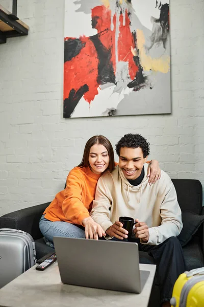 Sorrindo casal diversificado sentado confortavelmente em um sofá, posando com um laptop em um espaço de estar moderno — Fotografia de Stock