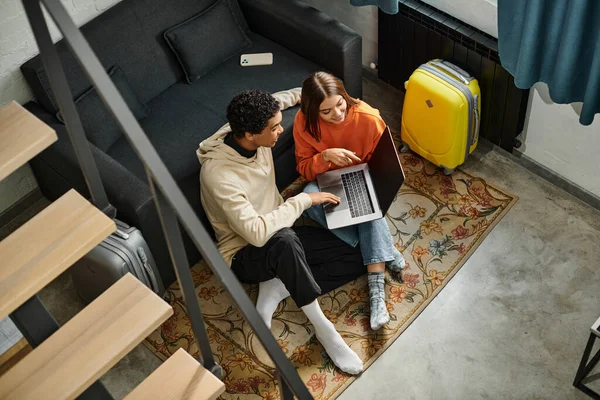 Diversa pareja mirando a un ordenador portátil mientras planea su próximo viaje, sentado en la alfombra cerca de las escaleras - foto de stock