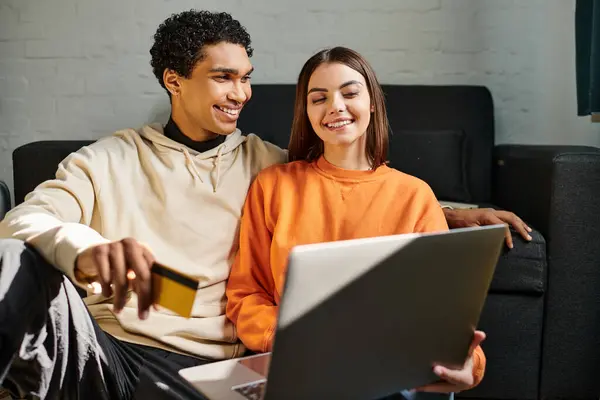 Diverso casal sorrindo compras on-line, segurando um cartão de crédito, com um laptop na sala de estar — Fotografia de Stock