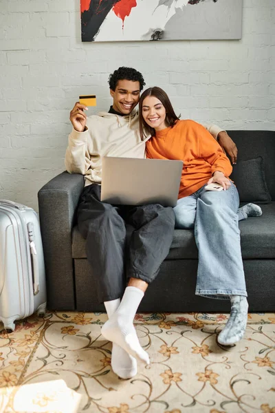 Jeune couple souriant magasinant en ligne, tenant une carte de crédit, avec un ordinateur portable sur leur canapé à la maison — Photo de stock