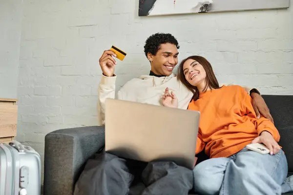 Heureux couple multiethnique shopping en ligne, tenant une carte de crédit, avec un ordinateur portable sur leur canapé à la maison — Photo de stock