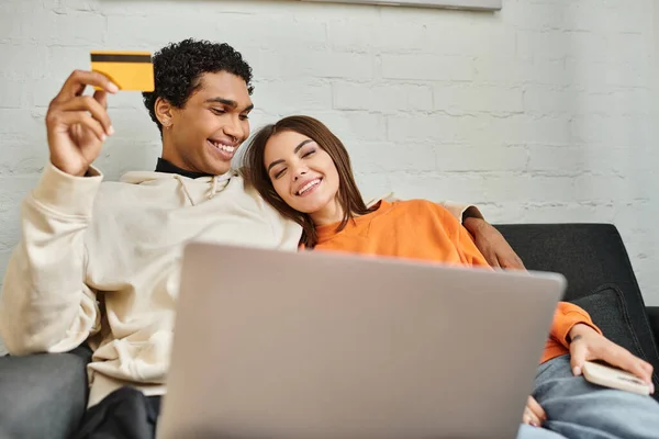 Feliz pareja multiétnica de compras en línea, con una tarjeta de crédito, con un ordenador portátil en el sofá gris en casa - foto de stock