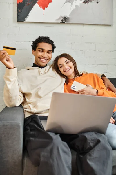 Feliz pareja multiétnica de compras en línea, con una tarjeta de crédito, con un ordenador portátil en el sofá en el albergue - foto de stock