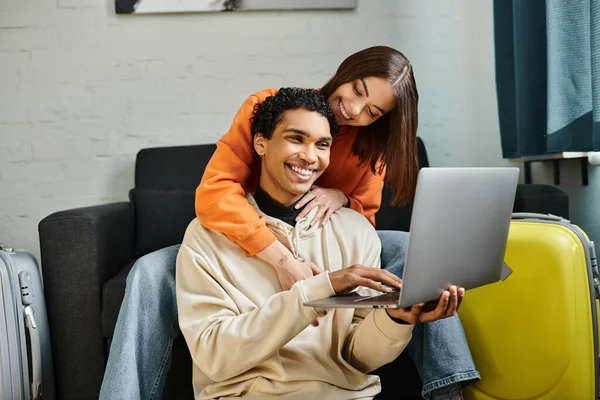 Alegre pareja multiétnica de compras en línea, la celebración de una tarjeta de crédito, con un ordenador portátil cerca de equipaje - foto de stock