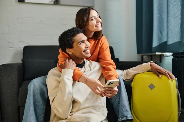 Happy woman embraces her black boyfriend on a cozy couch and holding smartphone near him, travel — Stock Photo