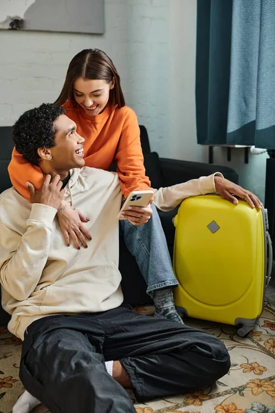 Happy woman holding smartphone and hugging african american boyfriend near luggage in hostel room — Stock Photo