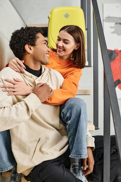 Positive diverse couple sitting on stairs and smiling at each other during their trip, hostel room — Stock Photo