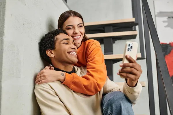 Glückliches Paar, das auf der Treppe sitzt und lächelt, während es im Hostel ein Selfie mit dem Smartphone macht — Stockfoto
