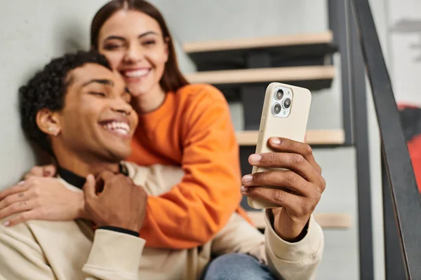 Alegre casal diversificado sentado nas escadas e sorrindo ao tomar selfie no smartphone no albergue — Fotografia de Stock