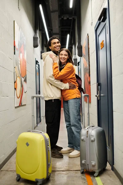 Feliz pareja diversa abrazándose en la sala del albergue con maletas, disfrutando de una experiencia de viaje alegre - foto de stock