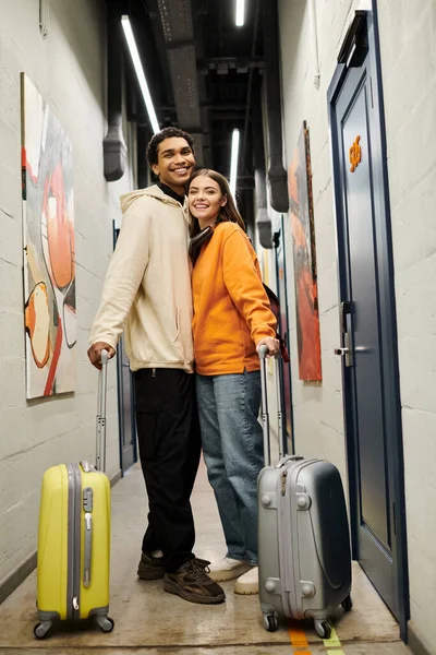 Feliz pareja diversa en un corredor de albergue con maletas, disfrutando de una experiencia de viaje alegre - foto de stock
