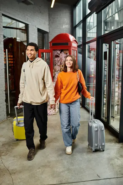 Interracial feliz pareja camina a través de un pasillo cogido de la mano y tirando de una maleta, viaje - foto de stock