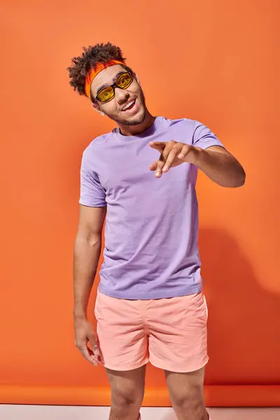 Feliz joven afroamericano hombre en gafas apuntando con el dedo sobre fondo naranja, gesto - foto de stock