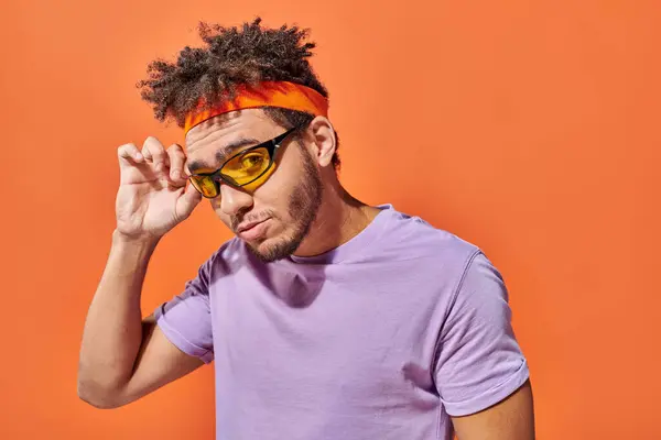 Handsome young african american man adjusting sunglasses and looking at camera on orange background — Stock Photo