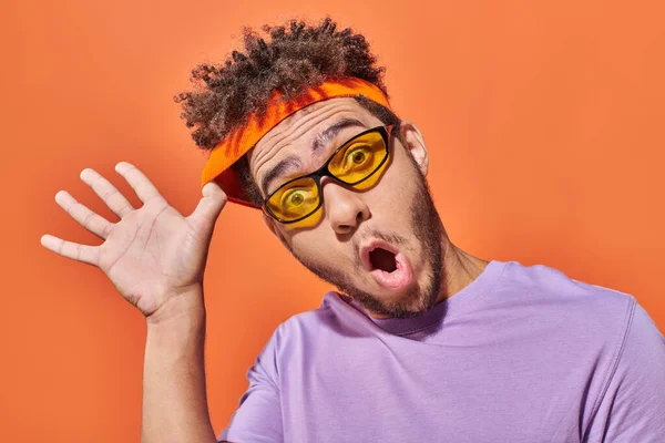 Funny african american man in sunglasses adjusting headband on orange background, surprised face — Stock Photo