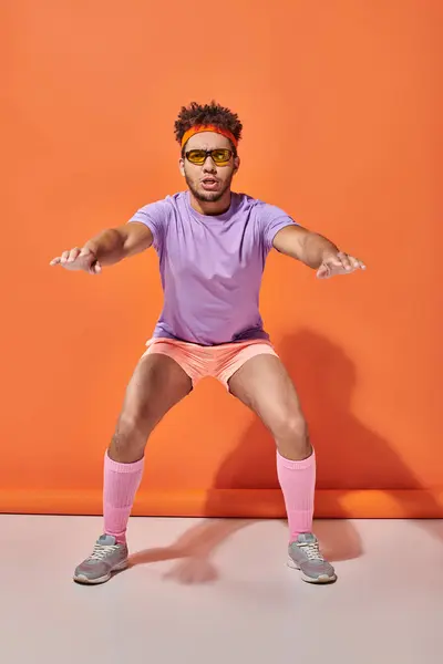 Sportive african american man in sunglasses and gym attire squatting on orange background — Stock Photo