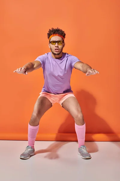 Deportivo afroamericano hombre en gafas de sol y traje de gimnasio en cuclillas sobre fondo naranja - foto de stock
