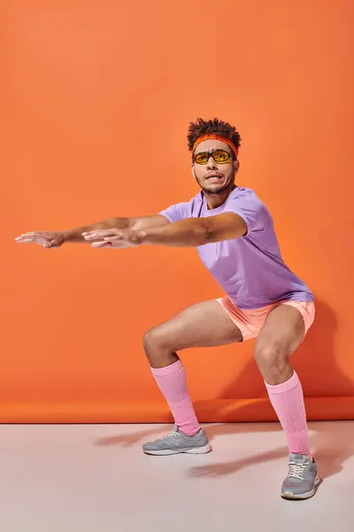 Young african american sportsman in sunglasses and gym attire squatting on orange background — Stock Photo