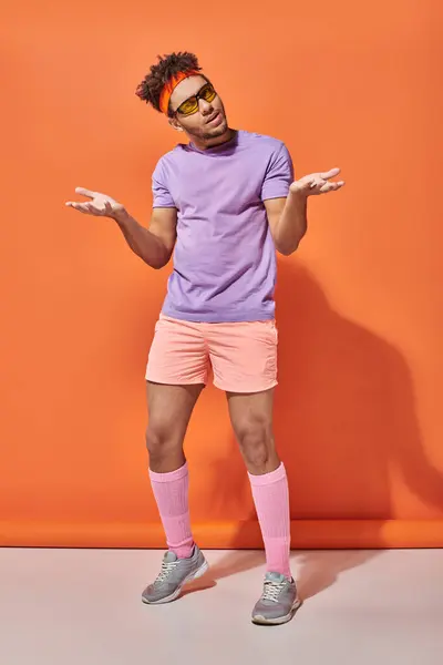 Confused african american sportsman in sunglasses and gym attire gesturing on orange background — Stock Photo