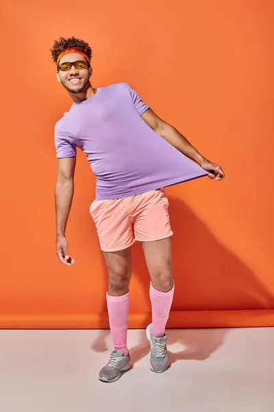 Alegre afroamericano deportista en traje de gimnasio mostrando su camiseta púrpura sobre fondo naranja - foto de stock