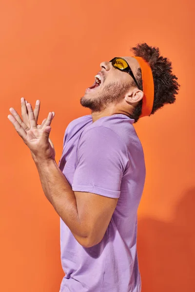 Expressive african american man in eyeglasses and headband screaming on orange background — Stock Photo