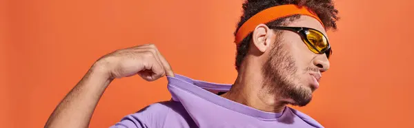 Banner of confident african american man in headband adjusting purple t-shirt on orange background — Stock Photo