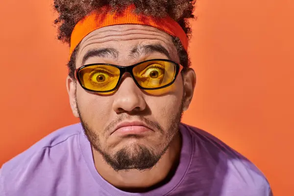 Sad african american man in eyeglasses and headband grinning on orange background, grimace — Stock Photo