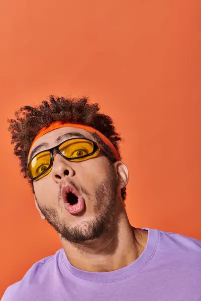 Homme afro-américain choqué dans les lunettes et bandeau regardant caméra sur fond orange — Photo de stock