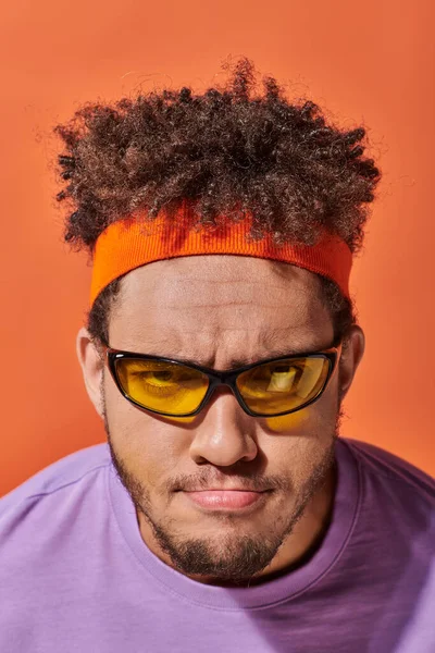 Jeune homme afro-américain en lunettes et bandeau regardant la caméra sur fond orange, fronçant les sourcils — Photo de stock