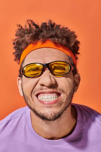Drôle afro-américain gars dans les lunettes et bandeau sourire sur fond orange, grimace — Photo de stock